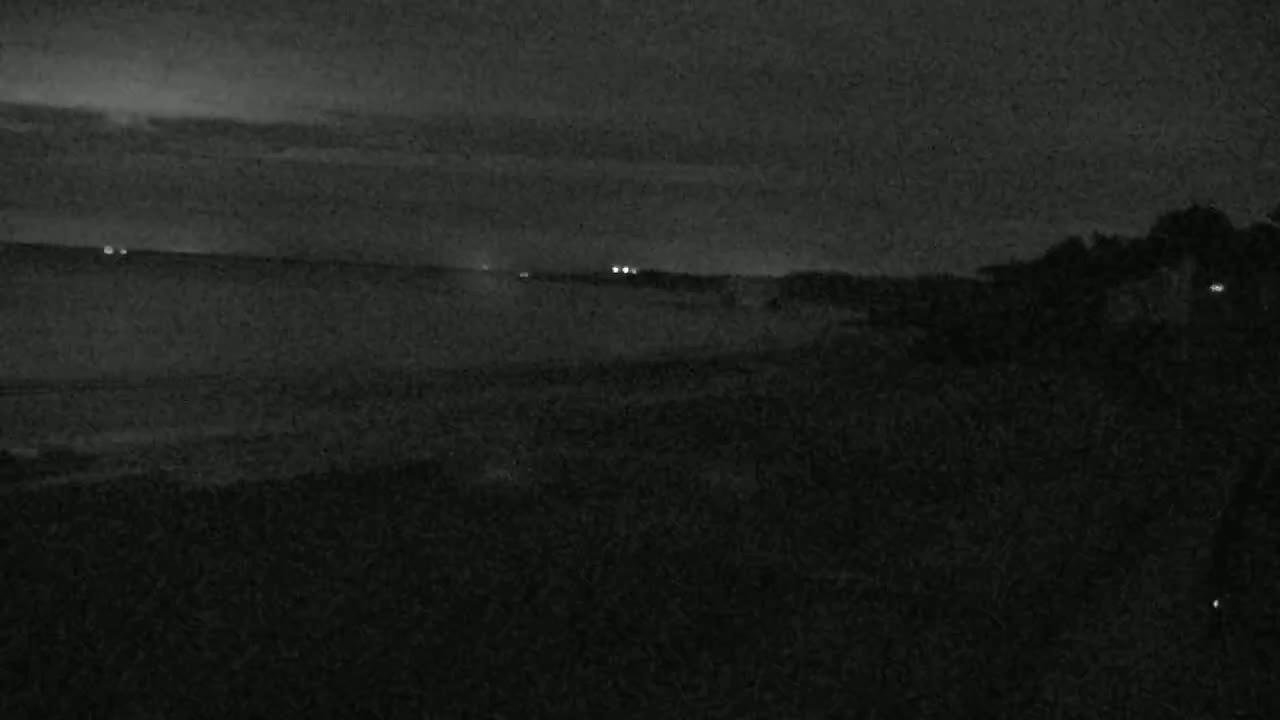 A dark, nighttime scene shows a calm body of water under a mostly cloudy sky, with some vegetation visible in the foreground and a few distant lights.
