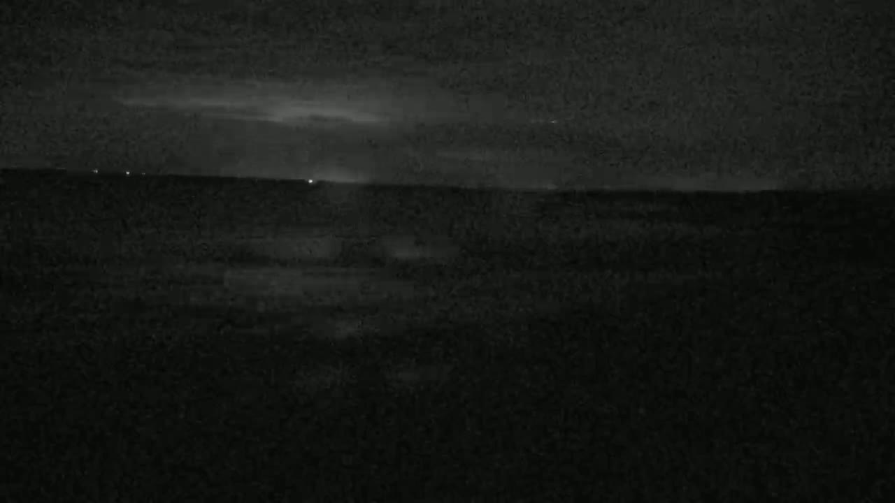 A dark, nighttime scene shows a flat, dark expanse of land or water under a cloudy sky with faint lights visible on the horizon.