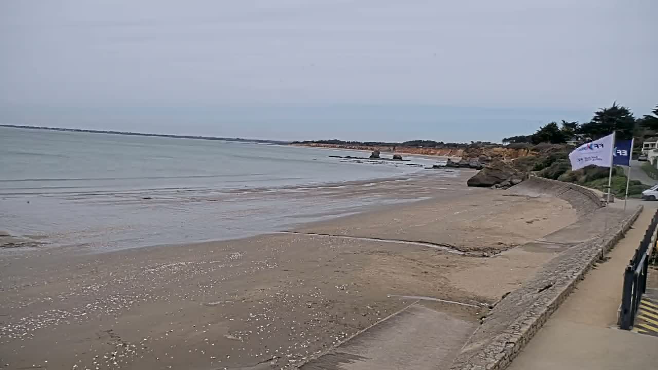 A sandy beach stretches along a calm, gray sea under an overcast sky, with a low stone wall separating the beach from a paved walkway and a few trees in the distance.