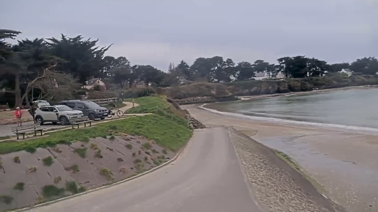 A paved road leads down to a sandy beach with gentle waves on an overcast day, with several cars parked along a grassy area beside it, near some trees.