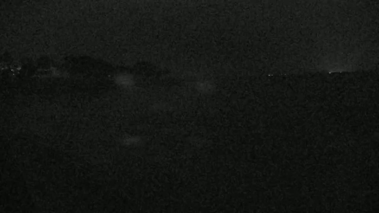 A dark, nighttime scene shows a distant, dimly lit shoreline under a cloudy sky.