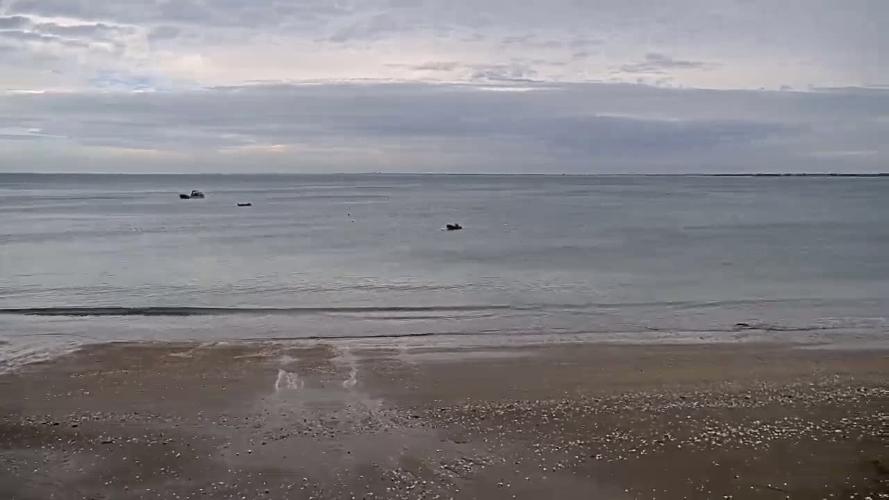 A sandy beach with small waves laps the shore under a cloudy sky, with a few small boats visible on the calm water in the distance.
