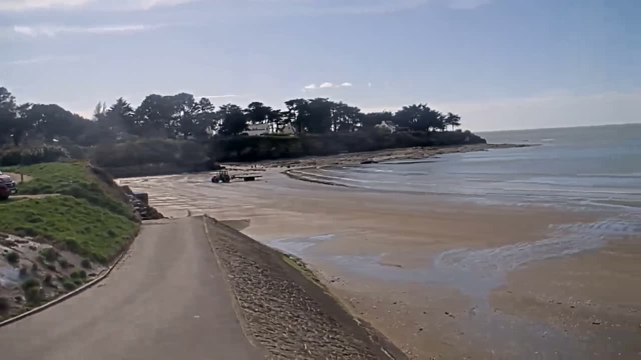 A sandy beach with calm water stretches out from a paved road leading to a line of trees and houses on a partly sunny day.