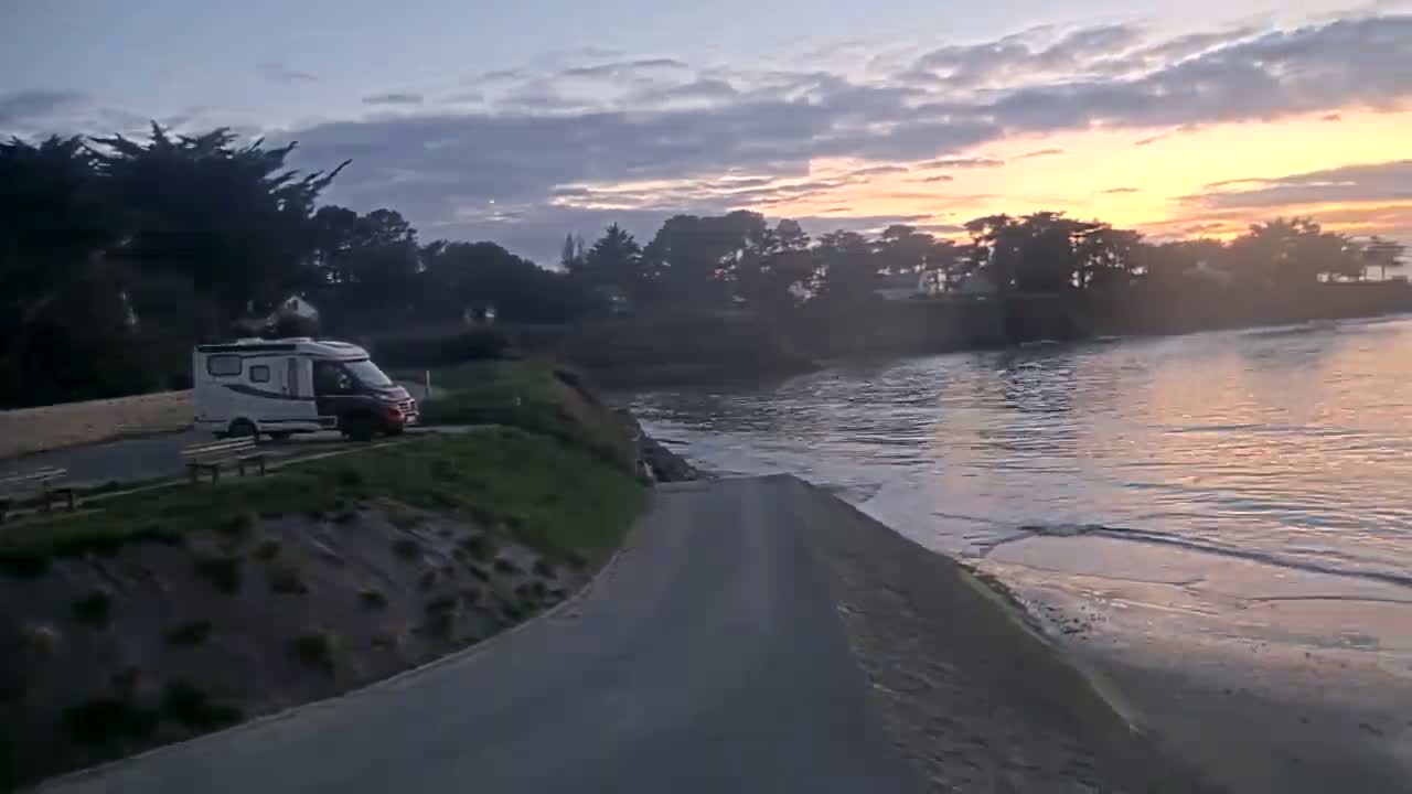 A campervan is parked near a calm body of water at sunset under a partly cloudy sky.