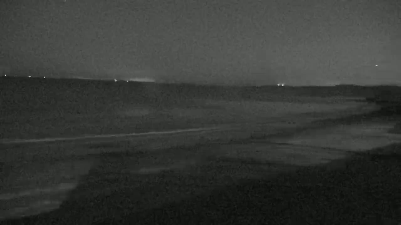 A dark, nighttime landscape shows a flat expanse of land with a faint horizon line and a few distant lights under a mostly clear sky.