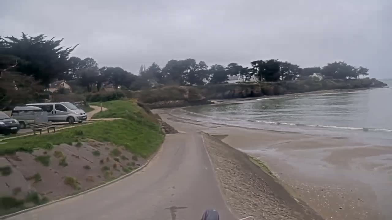 A paved road leads down to a sandy beach and calm ocean under an overcast sky, with several parked vans visible near some greenery and trees.