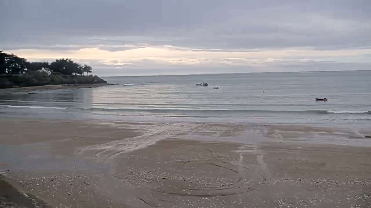 A sandy beach with gentle waves laps at the shore under a cloudy sky, with a few small boats visible in the calm water and trees on a small headland in the background.