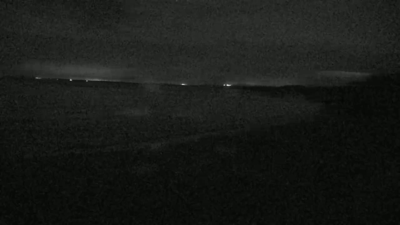A dark, nighttime landscape with a few distant lights on the horizon under a cloudy sky.