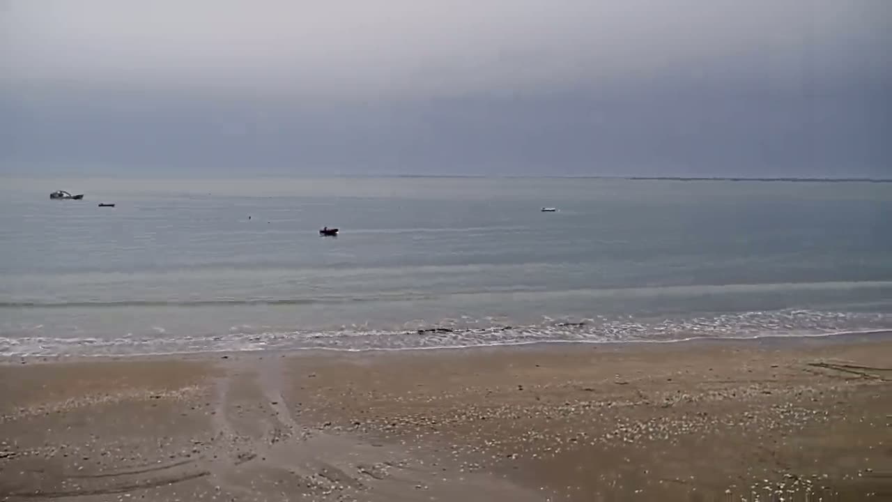 A sandy beach with gentle waves laps the shore under an overcast sky, with several small boats visible in the calm sea.
