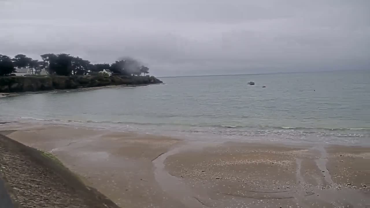 A sandy beach with calm, pale-green water under an overcast sky stretches towards a line of trees and houses on a distant headland.