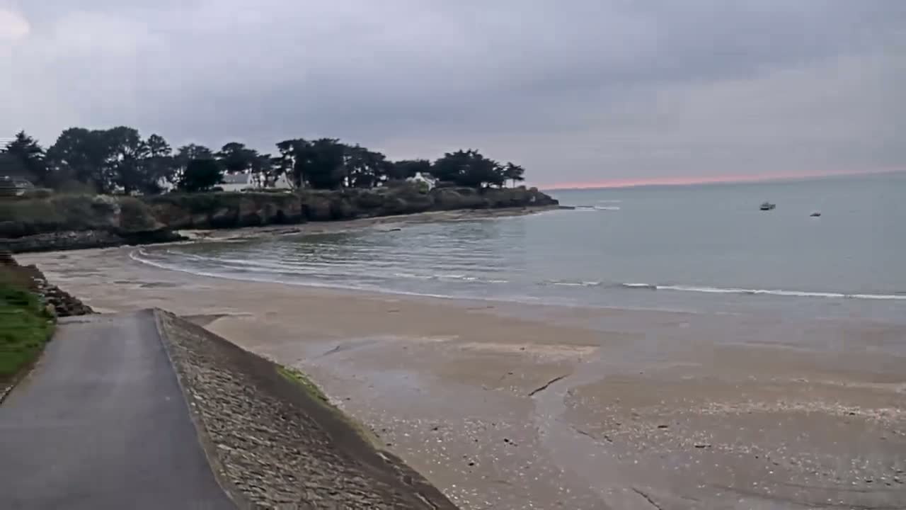 A sandy beach curves into a calm, light-grey sea under an overcast sky, with a paved road leading down to the water's edge and trees and houses visible on a cliff in the background.