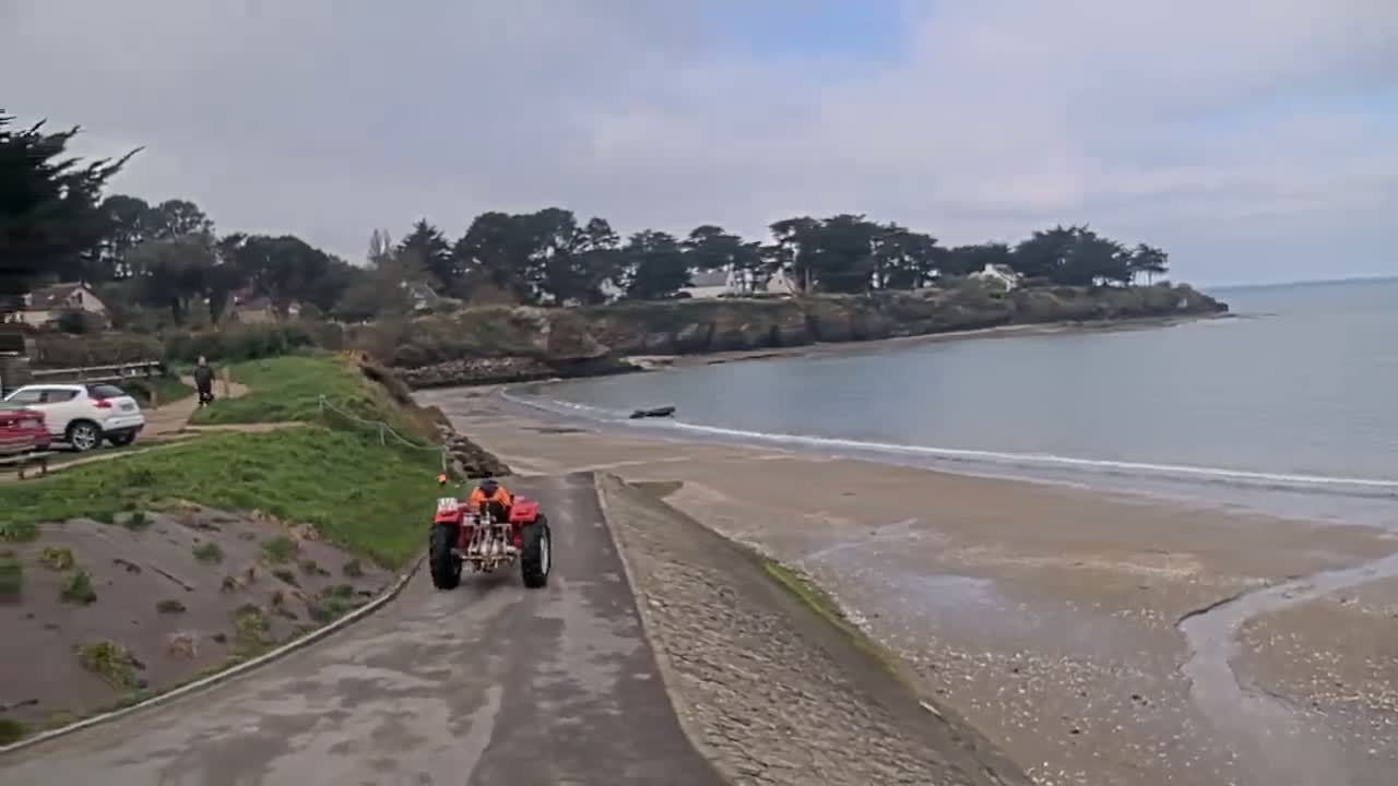 A red utility vehicle drives on a paved road overlooking a sandy beach and calm bay under an overcast sky.