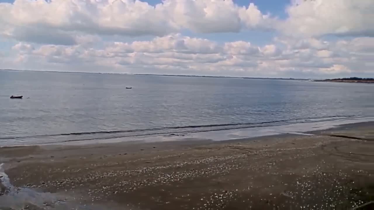 A sandy beach stretches towards a calm sea under a partly cloudy sky, with a few small boats visible on the water.