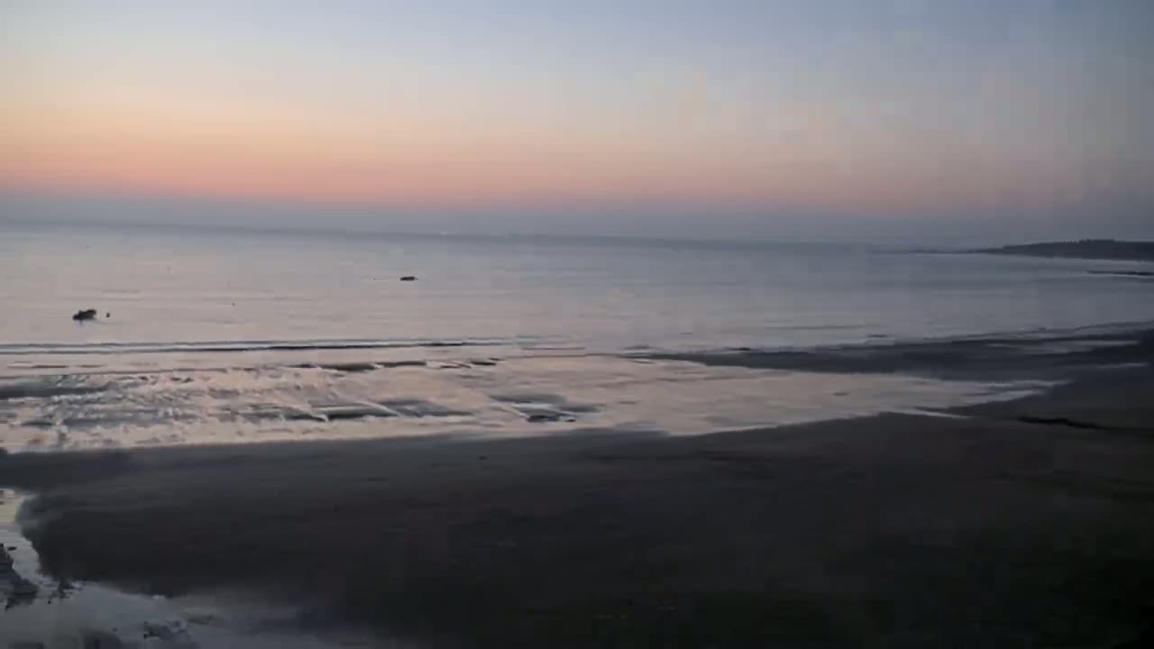 A calm ocean at sunset reveals a wide, dark sandy beach at low tide.