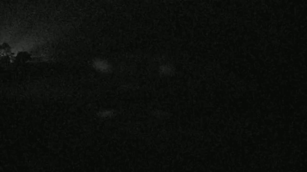A dark, nighttime image shows a mostly obscured landscape with faint, blurry light sources in the distance under a cloudy sky.