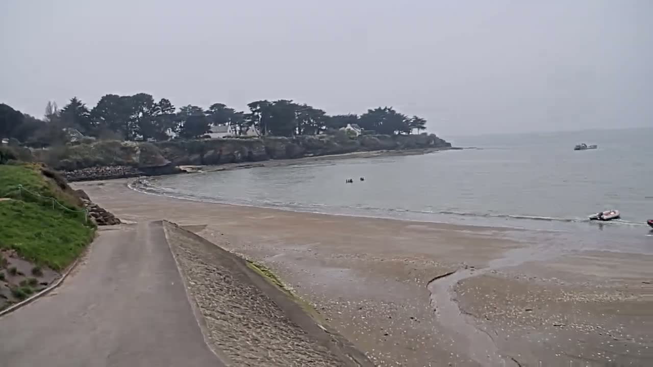 A sandy beach and calm, gray water are visible under an overcast sky, with a road leading down to the shore from a grassy area, and a few small boats and people in the water.