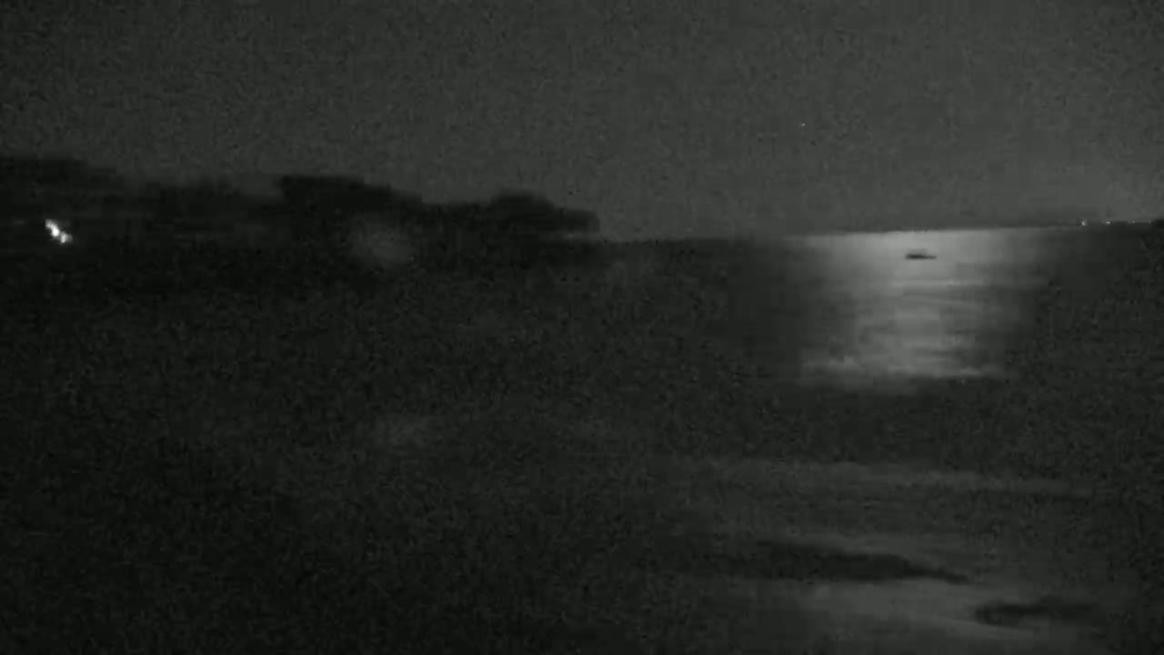 A dark, nighttime scene shows a body of water reflecting light, with a small vessel visible on the water's surface.