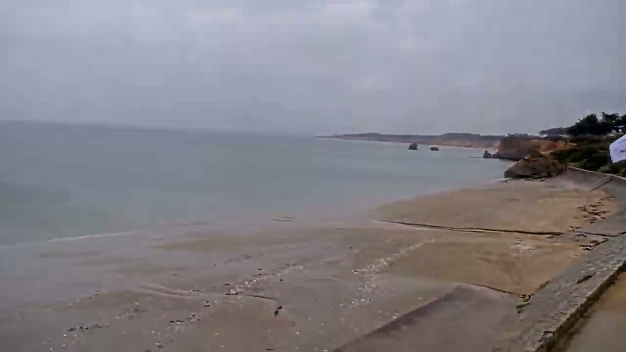 A flat, sandy beach stretches to a calm, grey sea under an overcast sky, with rocky outcrops visible along the coastline.