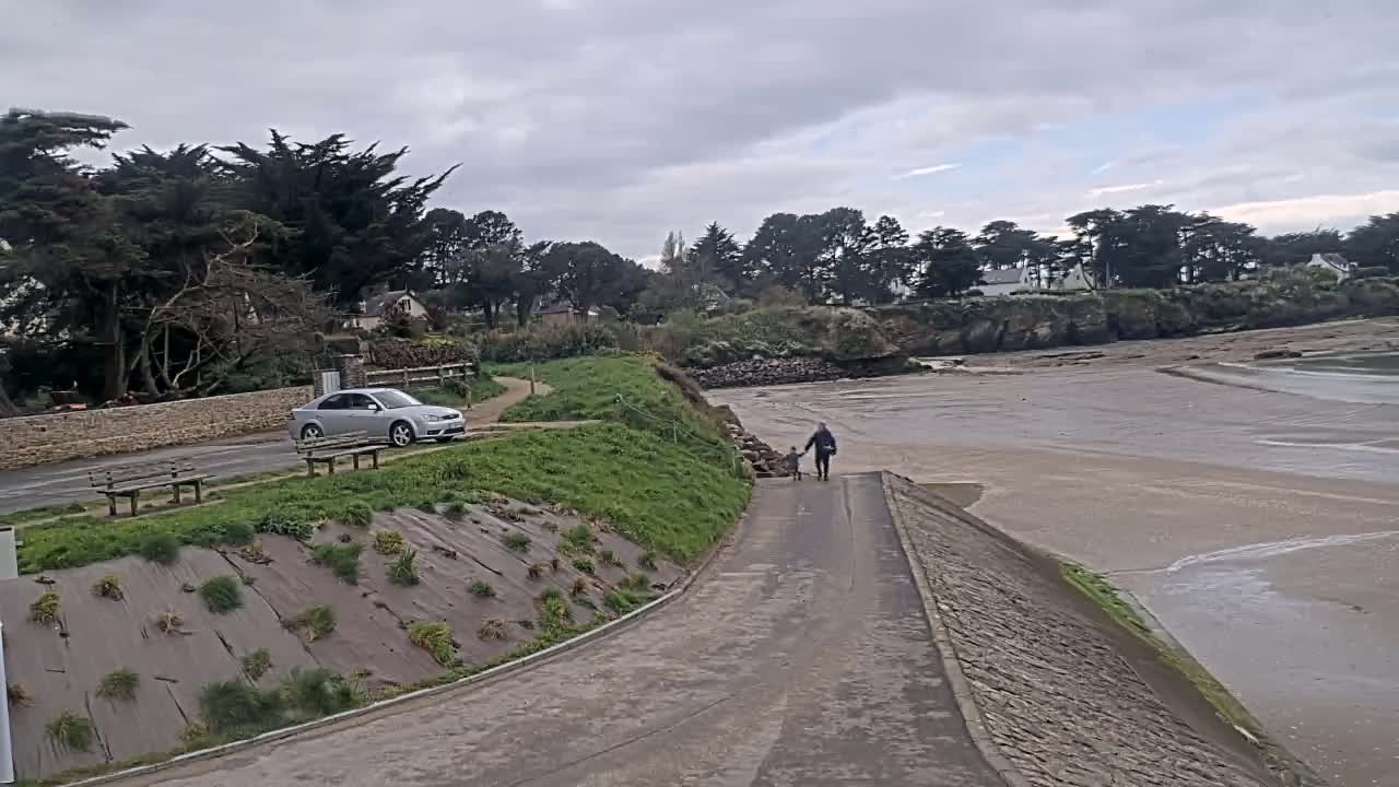 A paved path leads down to a sandy beach, where an adult and child walk, under an overcast sky.