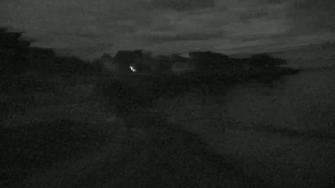 A dark, possibly nighttime, outdoor scene shows a small, bright light in the middle distance against a mostly featureless, dark landscape under a cloudy sky.