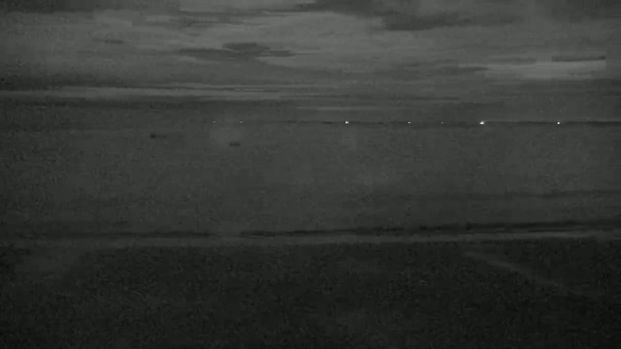 A dark, overcast sky looms over a calm body of water, with a few faint lights visible on the horizon.