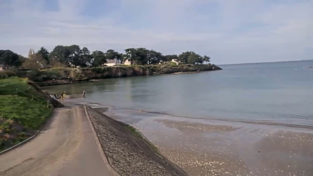 A paved road leads down to a calm bay under a partly cloudy sky, where several people are standing near the water's edge.