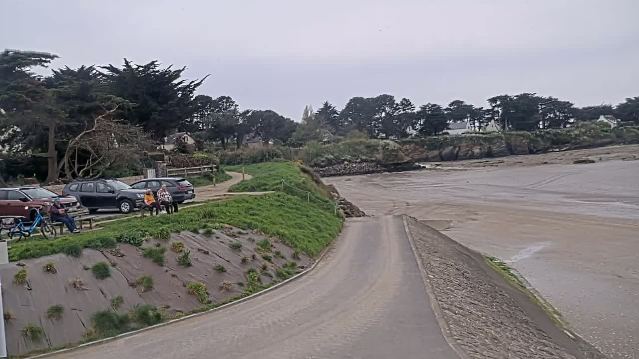 A paved road leads down to a sandy beach, with several cars parked and people sitting near some trees under an overcast sky.