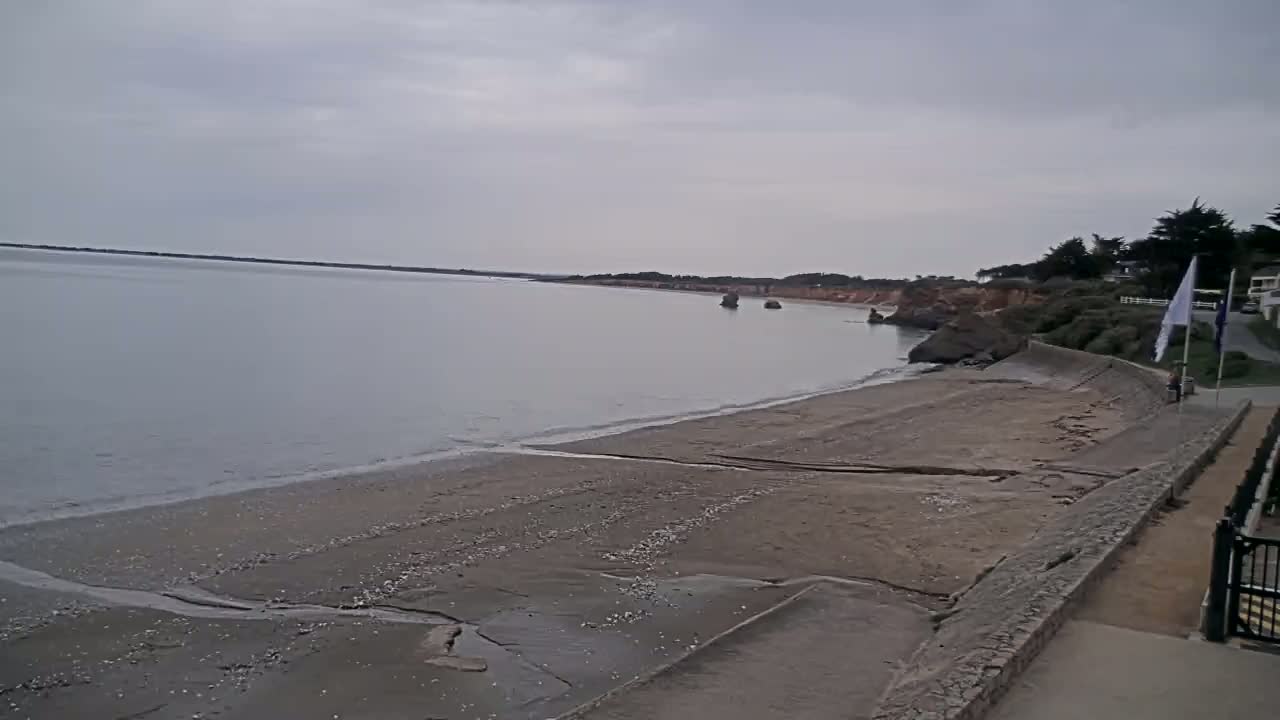 A calm, gray day reveals a sandy beach with a low sea wall, bordering a body of water, and a distant shoreline with some vegetation.