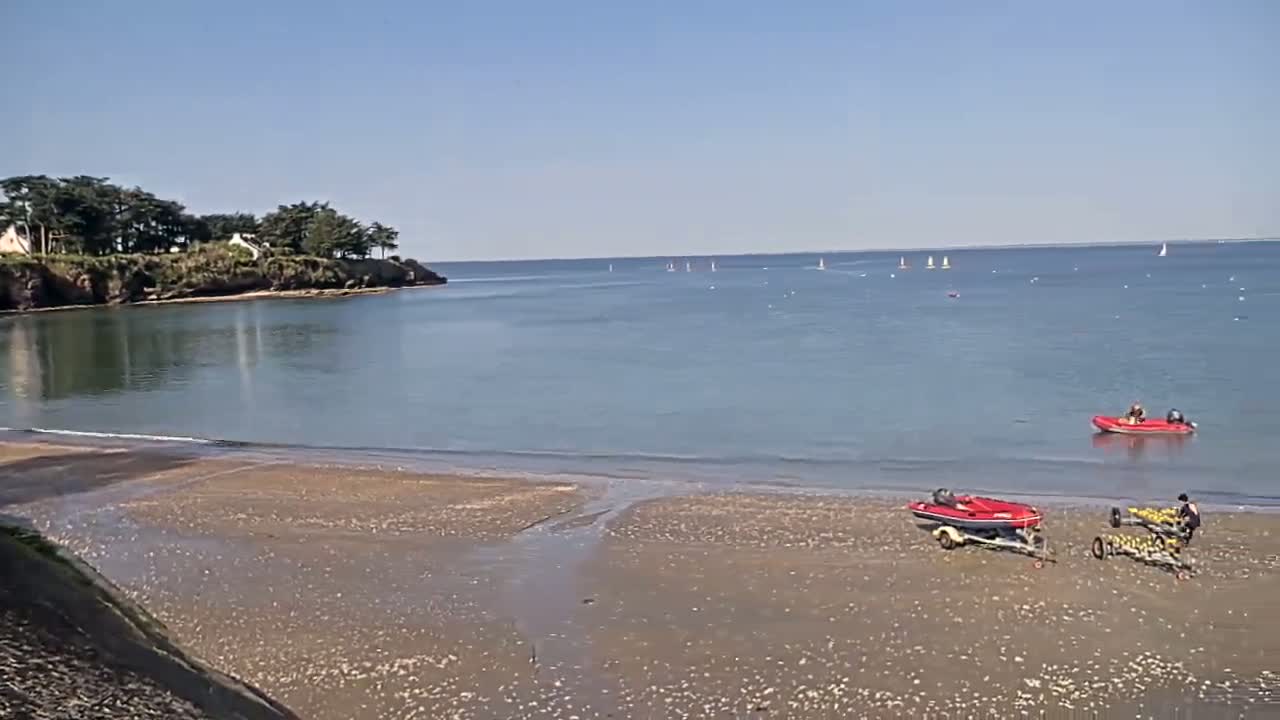 A sandy beach with several small boats on it and in the water is nestled in a calm bay under a clear blue sky, with a small, tree-covered landmass visible in the distance.