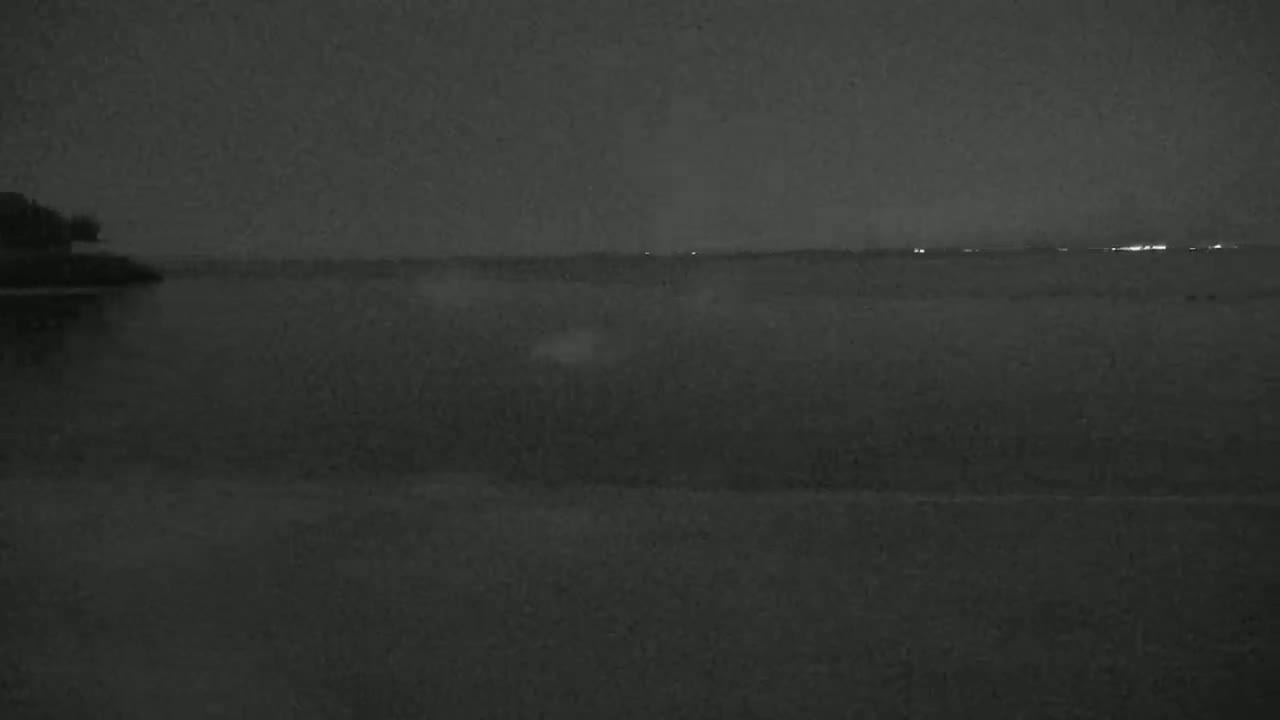 A dark, nighttime scene shows a body of water with a distant shoreline faintly lit by lights, under a mostly cloudy sky.