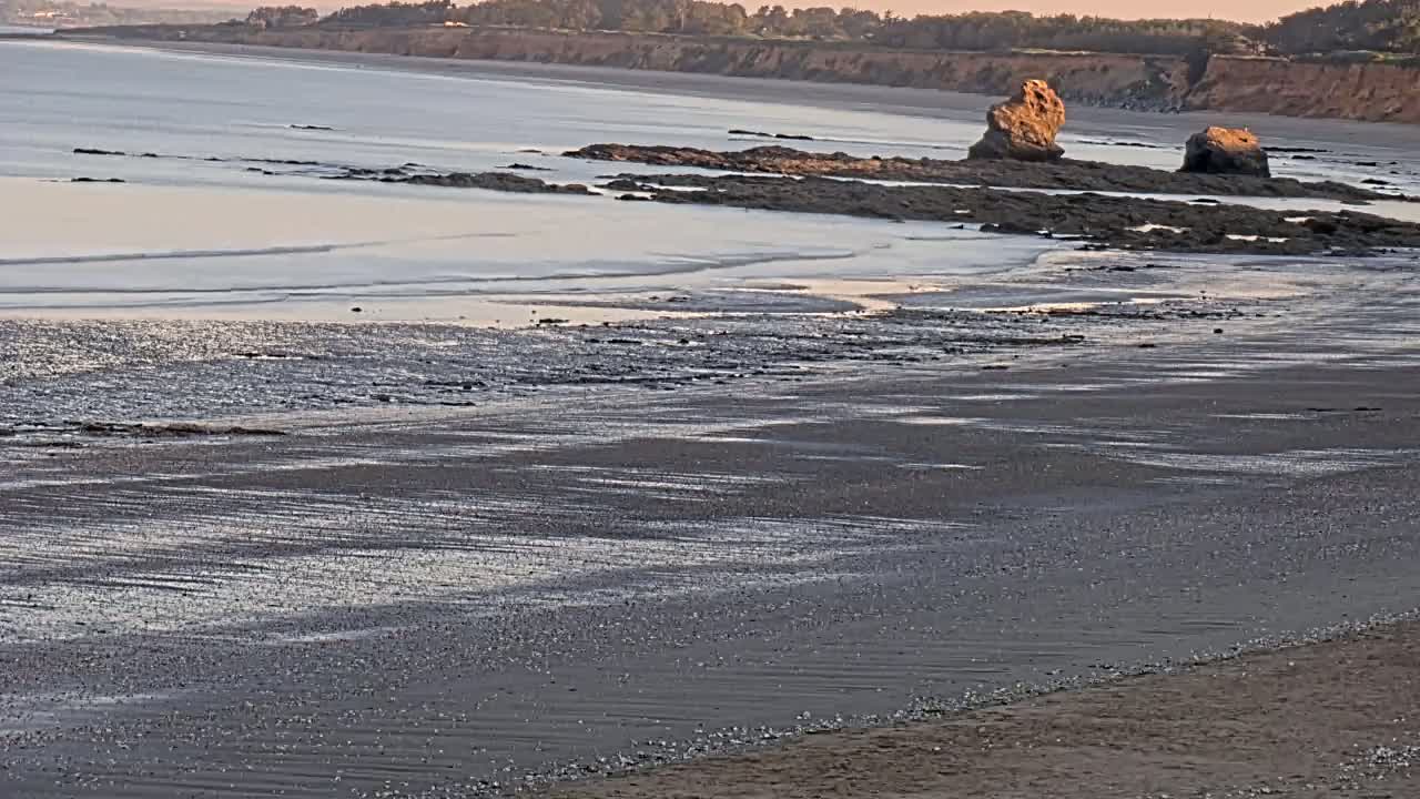 A calm, sandy beach stretches out to a calm ocean with dark rocks and cliffs in the background.