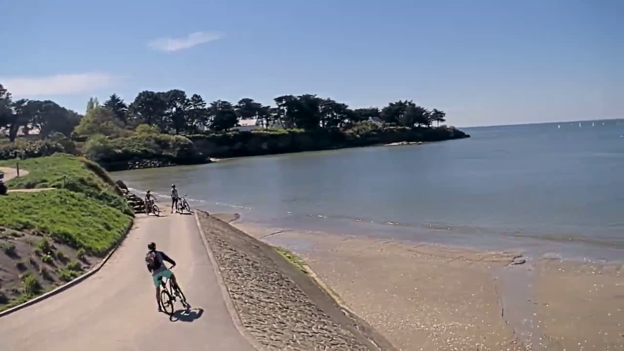Several cyclists ride along a paved path next to a calm body of water on a sunny day.