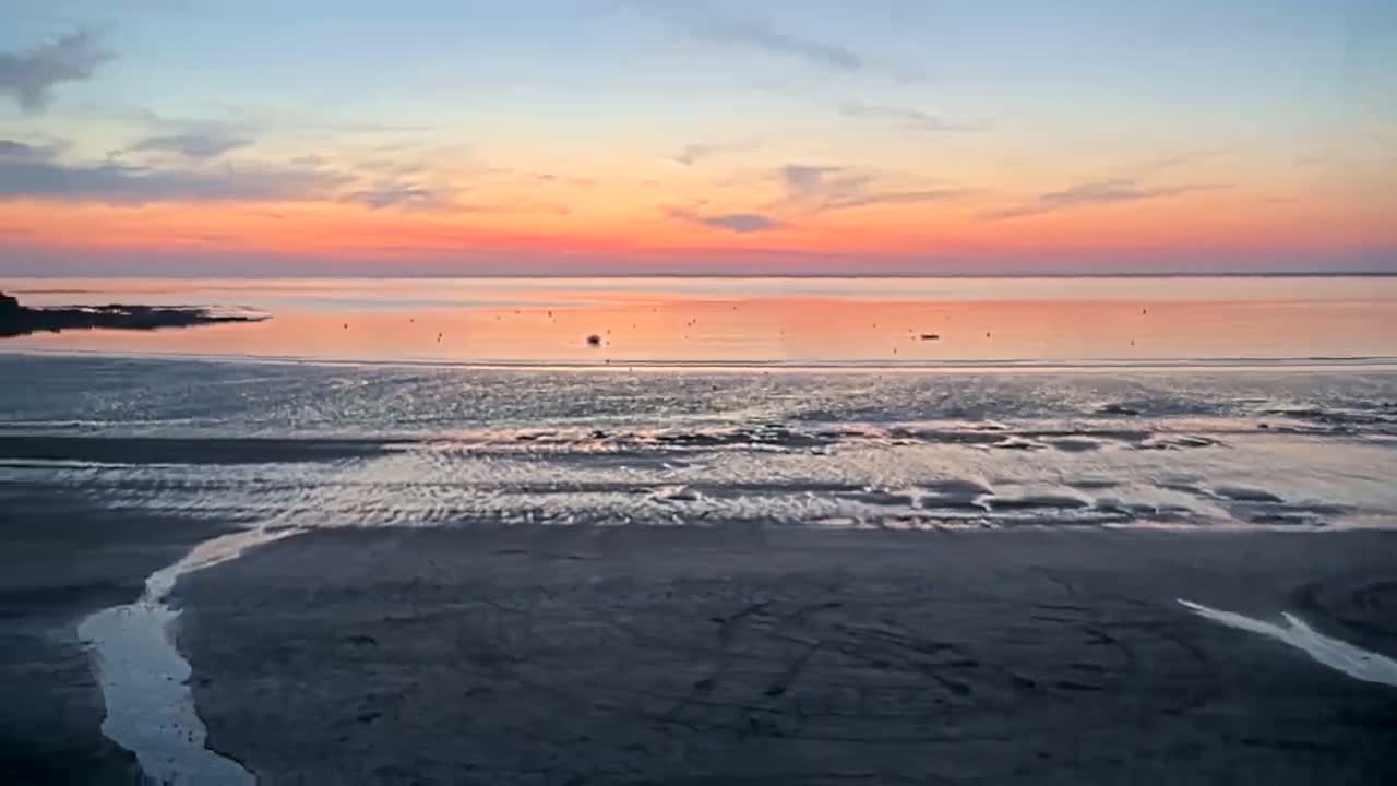 A pink and orange sunset over a calm ocean, reflecting on a wet, sandy beach at low tide.