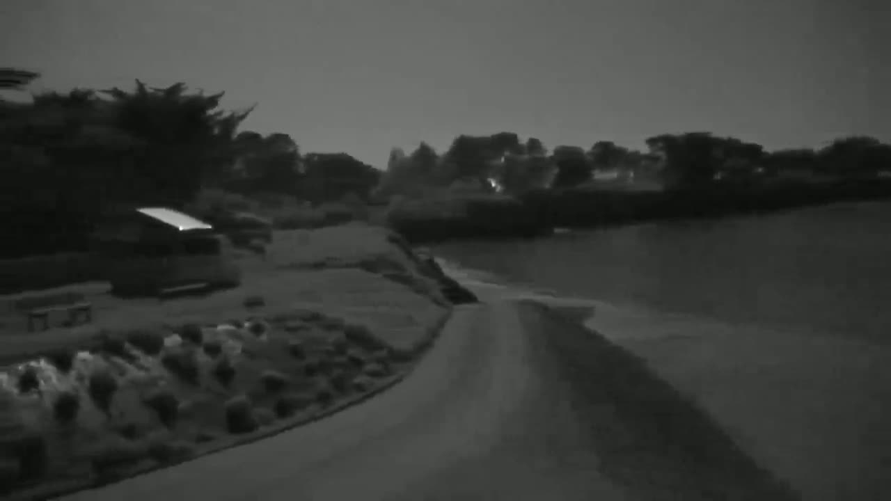 A nighttime, grayscale image shows a paved road curving toward a body of water, bordered by vegetation and a parked vehicle.