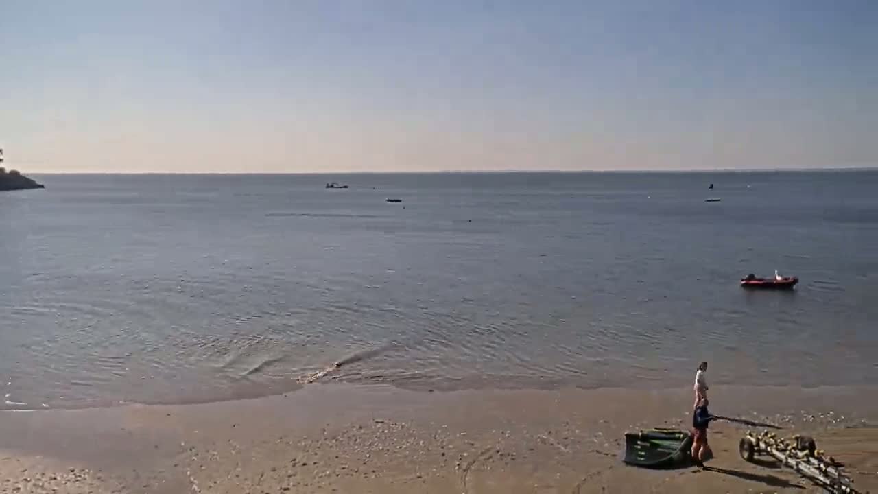 A sandy beach meets calm, clear water under a bright, sunny sky; a person stands on the beach near a small boat and a boat trailer.