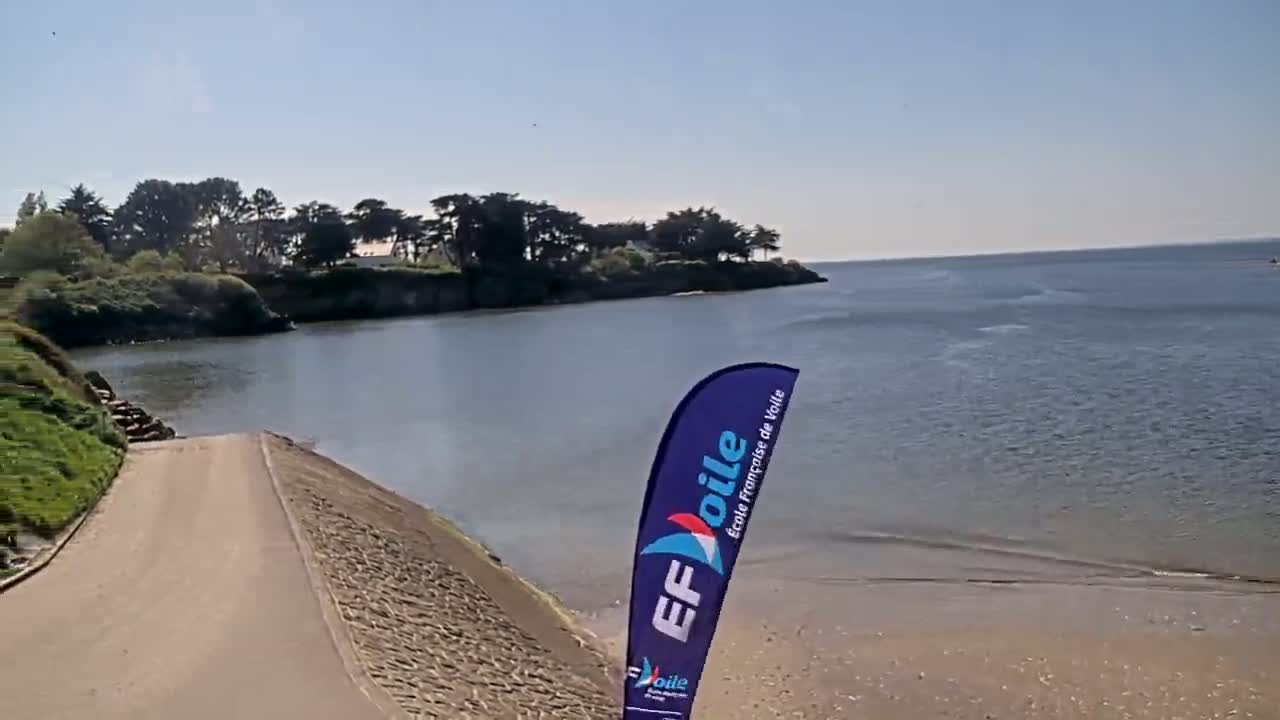A paved ramp leads down to a sandy beach beside a calm body of water under a clear, sunny sky, with a line of trees visible in the distance.