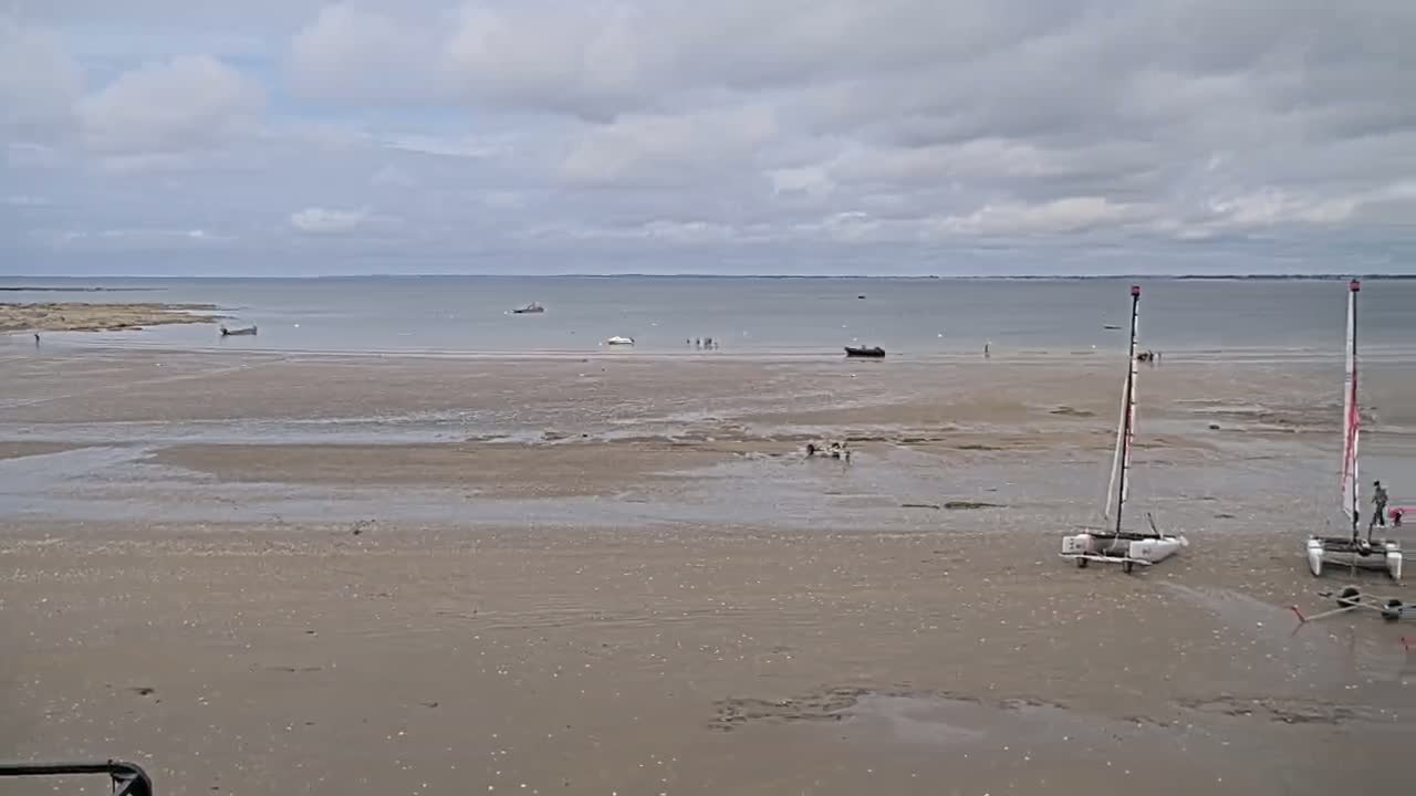 A wide expanse of sandy beach at low tide stretches towards a calm sea under a cloudy sky, with several small boats and two catamarans grounded on the sand.