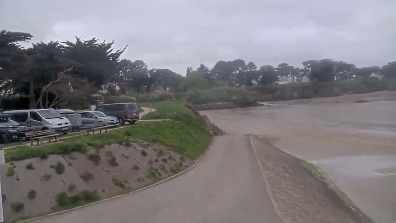 A paved road curves down to a muddy, flat expanse of water under an overcast sky, with several cars parked in a lot beside some green vegetation.