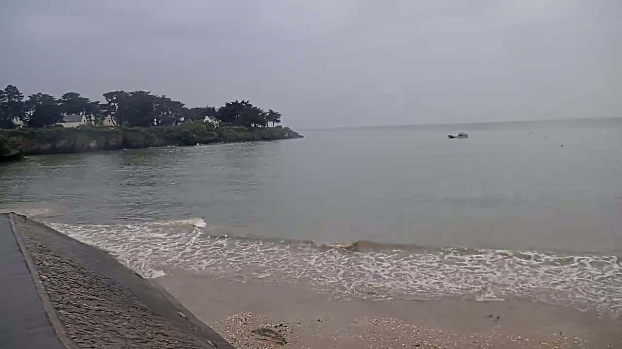 A sandy beach with small waves laps against a sea wall under an overcast sky, with a small boat visible in the calm water and a line of trees and houses on a distant headland.