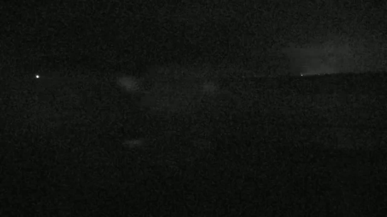 A nighttime, low-light view of a dark landscape with a few faint lights visible in the distance under a mostly cloudy sky.