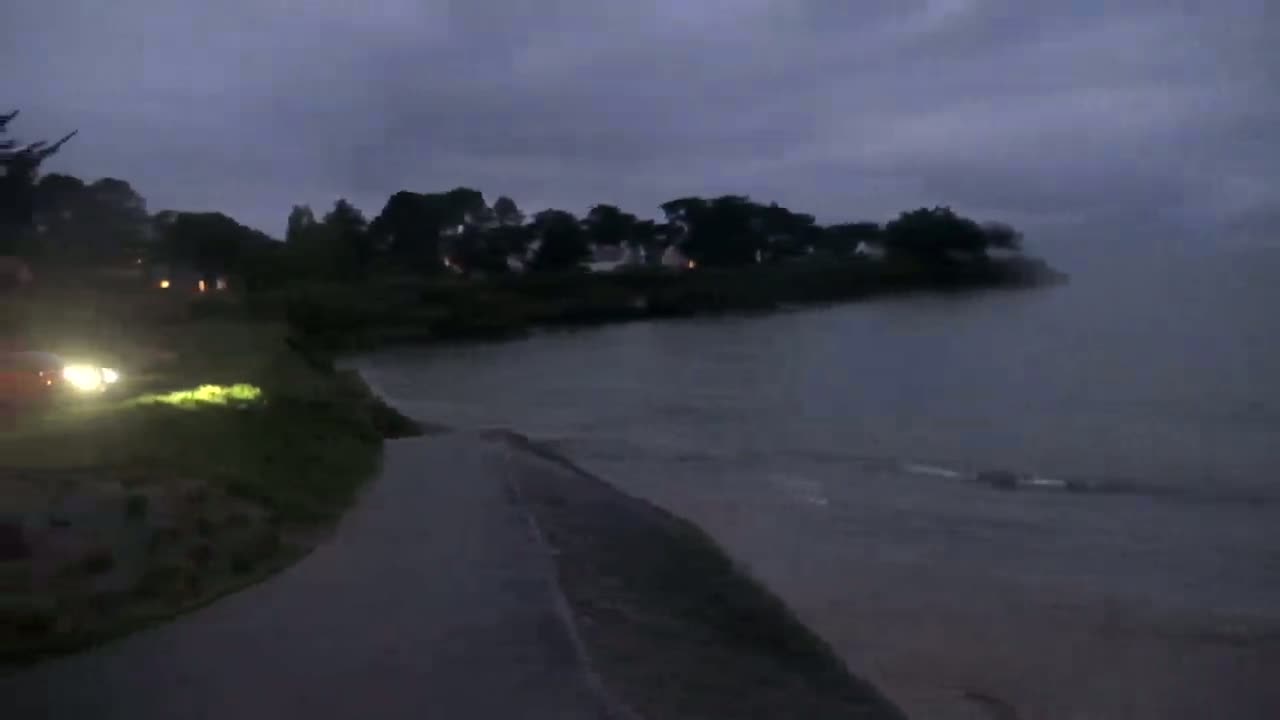 A dark, overcast night scene shows a body of water next to a paved path and a line of trees and houses.