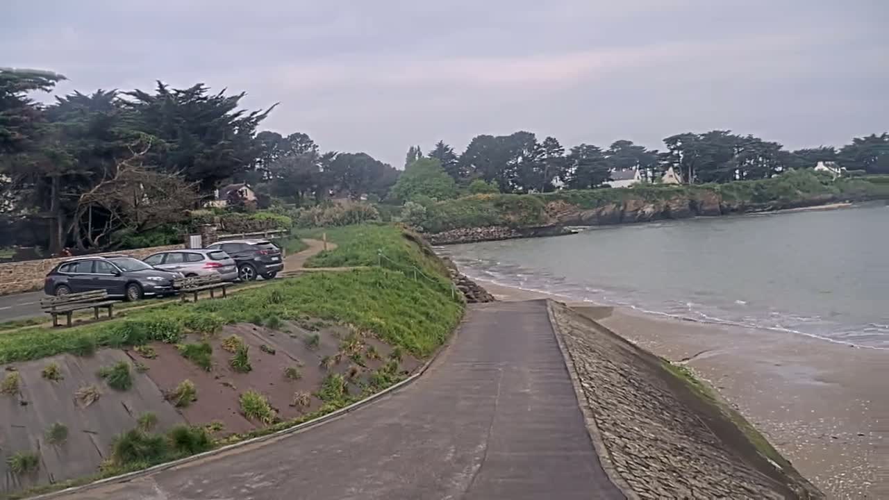 A paved road leads down to a sandy beach with calm waves on a cloudy day, with several cars parked near some trees and houses in the background.