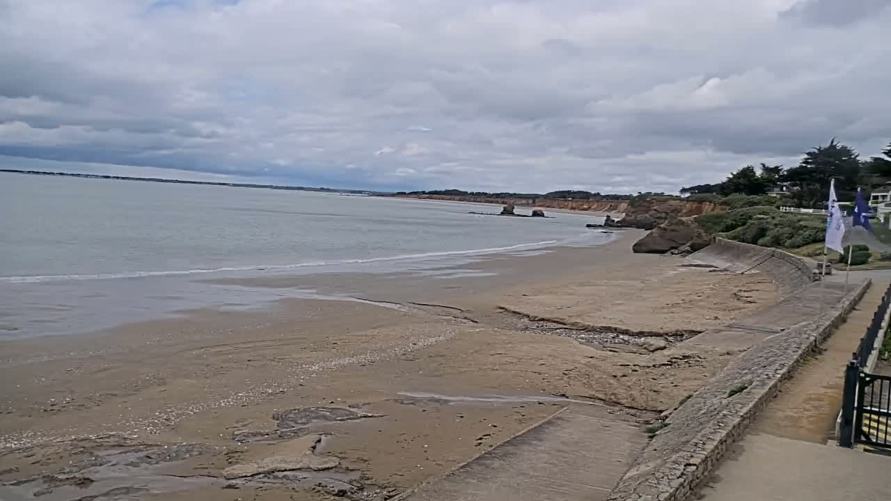 A sandy beach with a calm ocean under a cloudy sky is seen from a high vantage point, with a stone wall and vegetation bordering the beach.