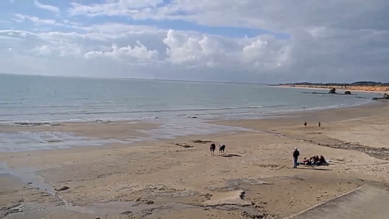 A sandy beach stretches out to a calm sea under a partly cloudy sky, with a few people scattered along the shore.