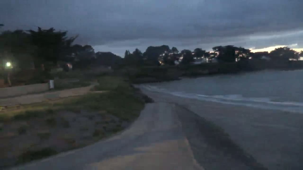A paved path leads down to a beach at dusk under a cloudy sky, with calm waves lapping the shore.