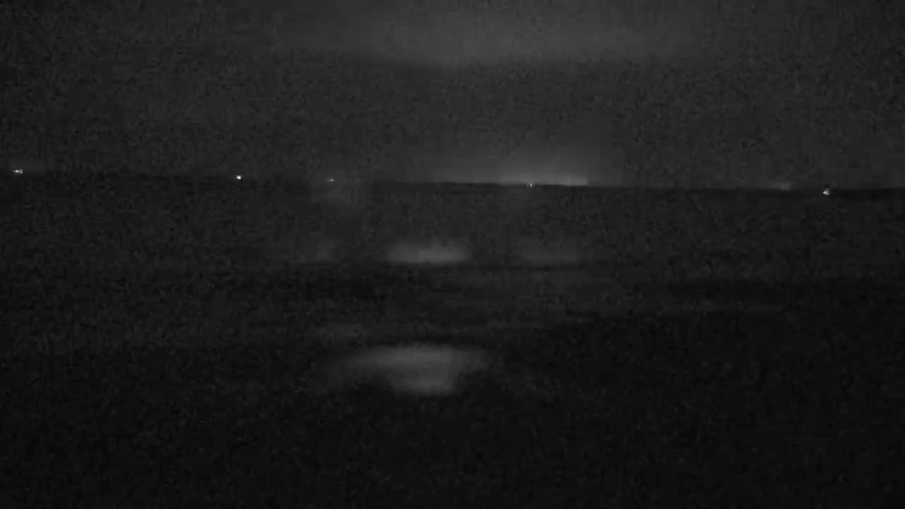 A nighttime, dark, and possibly stormy outdoor scene shows a flat expanse of land with a few faint lights in the distance and dark, slightly reflective puddles in the foreground.