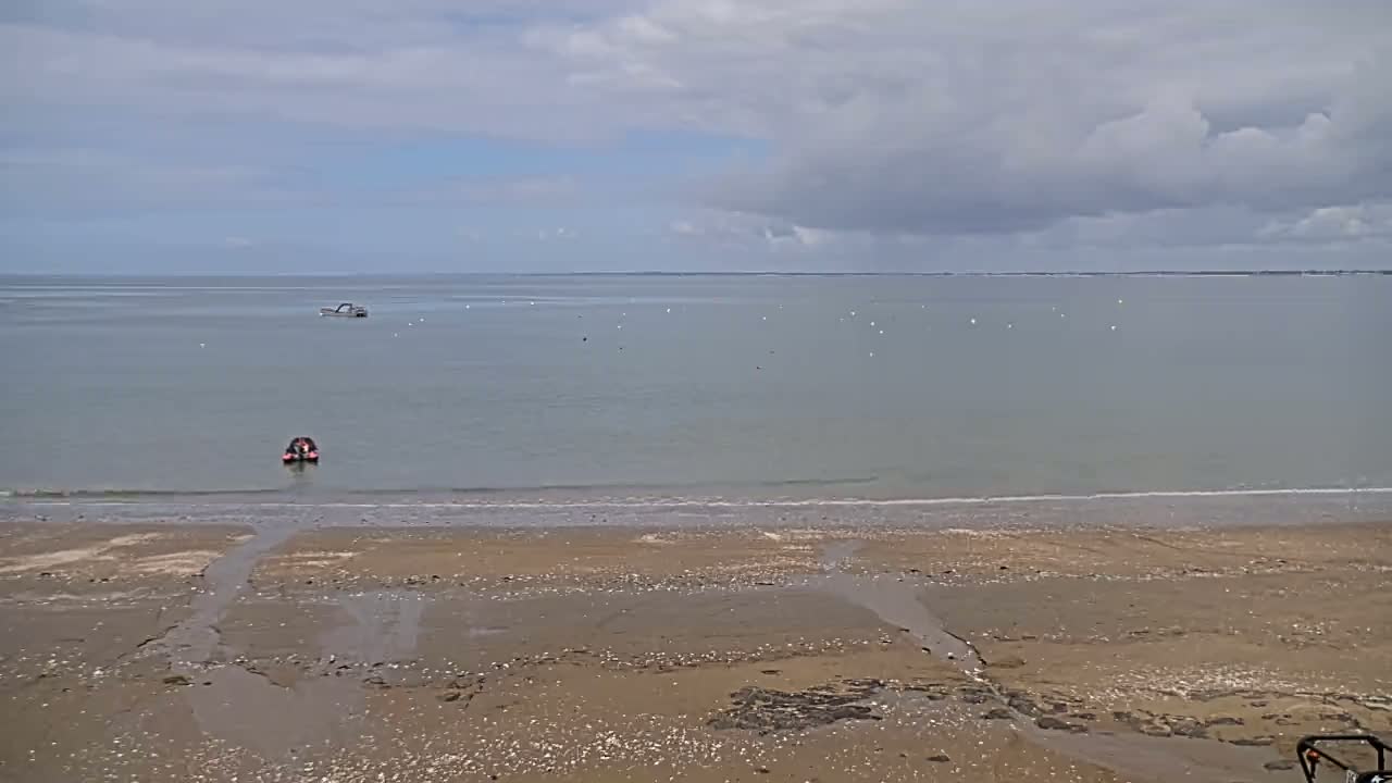 A sandy beach stretches to a calm, gray-green sea under a mostly cloudy sky, with a few small boats visible on the water.