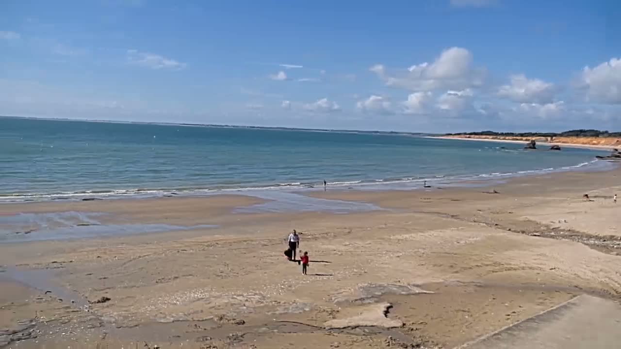 A wide sandy beach stretches to a calm ocean under a mostly sunny sky with a few scattered clouds, and two people and a child are visible walking on the beach.