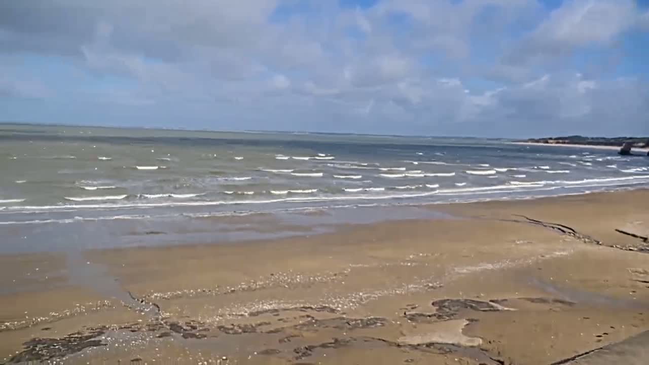 A wide sandy beach stretches to a moderately wavy sea under a partly cloudy sky.