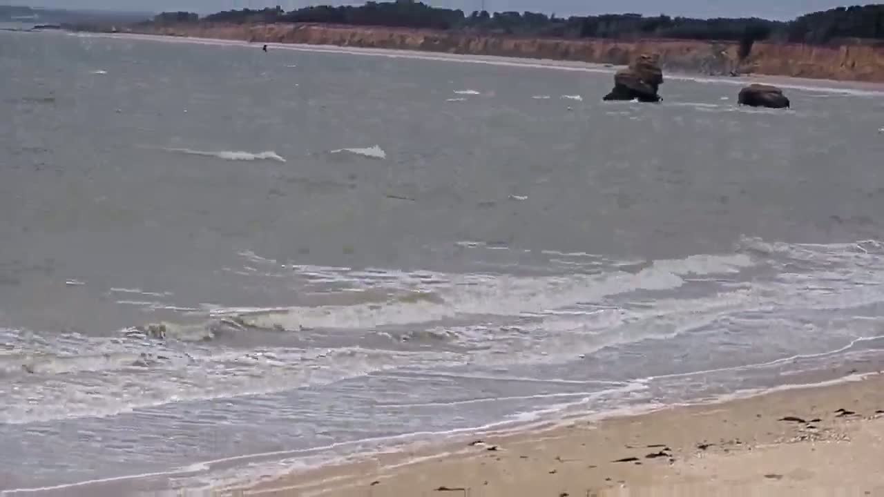 A sandy beach with small waves laps the shore under a cloudy sky, with a few rocks visible in the water and a distant shoreline.